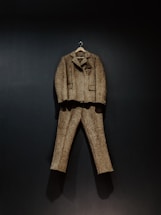 A brown tweed suit, consisting of a jacket and trousers, hangs against a dark background. The suit is displayed on a wooden hanger, showcasing its classic design and texture.