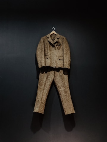 A brown tweed suit, consisting of a jacket and trousers, hangs against a dark background. The suit is displayed on a wooden hanger, showcasing its classic design and texture.