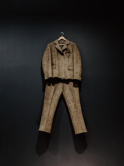 A brown tweed suit, consisting of a jacket and trousers, hangs against a dark background. The suit is displayed on a wooden hanger, showcasing its classic design and texture.