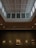 An art gallery hall with a high, coffered ceiling, illuminated by natural light from large windows. Several framed paintings are mounted on the dark walls, with varied artwork sizes and lighting that highlights their features.