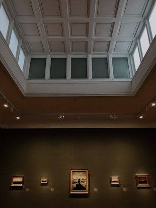An art gallery hall with a high, coffered ceiling, illuminated by natural light from large windows. Several framed paintings are mounted on the dark walls, with varied artwork sizes and lighting that highlights their features.