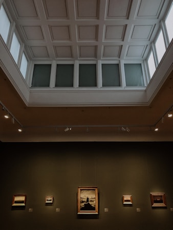 An art gallery hall with a high, coffered ceiling, illuminated by natural light from large windows. Several framed paintings are mounted on the dark walls, with varied artwork sizes and lighting that highlights their features.