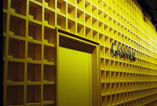 A wall with a grid pattern design painted in yellow with a doorway and the word 'CASHIER' prominently displayed on it in black letters.