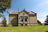 A large, traditional building constructed with light brown bricks, featuring prominent architectural elements such as a gabled roof and decorative stonework around the windows. The building is set against a backdrop of clear blue sky with a few trees on either side, surrounded by a well-maintained grassy lawn, suggesting a tranquil environment.