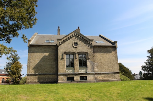 A large, traditional building constructed with light brown bricks, featuring prominent architectural elements such as a gabled roof and decorative stonework around the windows. The building is set against a backdrop of clear blue sky with a few trees on either side, surrounded by a well-maintained grassy lawn, suggesting a tranquil environment.