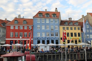 A vibrant row of colorful buildings with distinct architectural styles, featuring red, blue, and yellow facades and orange rooftops. The street level is bustling with people dining outdoors under white umbrellas, and several national flags are displayed. The scene exudes a lively and energetic atmosphere.