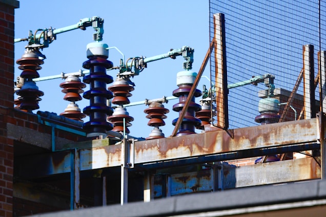 An array of large ceramic insulators with coiled structures are mounted on a rusty metal framework. The background includes a brick wall and a metal mesh fence, and the scene is set against a clear blue sky.