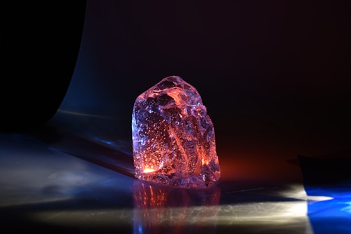 A translucent, irregularly shaped crystal or glass object sits on a reflective surface. It is illuminated by colorful lighting, primarily in shades of blue, orange, and pink, creating a glowing effect. The background is dark, enhancing the vibrant colors and the object's clarity.