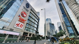 A bustling urban scene featuring a high-rise building with prominent retail brand logos such as CGV Cinemas and H&M. Other skyscrapers surround the area, with their reflective glass facades creating a modern city atmosphere. Palm trees line the streets, and a clear sky is visible above.