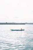 A lone fisherman wearing red sits in a small turquoise boat on a vast expanse of calm water. The background shows a distant shoreline with a few industrial structures and a hazy sky, creating a serene and peaceful atmosphere.