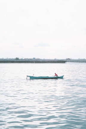 A lone fisherman wearing red sits in a small turquoise boat on a vast expanse of calm water. The background shows a distant shoreline with a few industrial structures and a hazy sky, creating a serene and peaceful atmosphere.