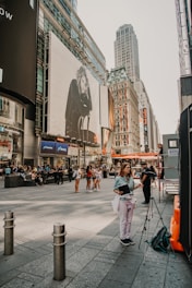 A bustling city street with people walking and gathering. Large billboards and tall buildings fill the scene, with one billboard featuring a monochrome image of a person sitting. A woman stands near a tripod with a bag and a notebook. The street scene is busy with various groups of people interacting and some individuals standing alone.