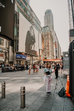 A bustling city street with people walking and gathering. Large billboards and tall buildings fill the scene, with one billboard featuring a monochrome image of a person sitting. A woman stands near a tripod with a bag and a notebook. The street scene is busy with various groups of people interacting and some individuals standing alone.