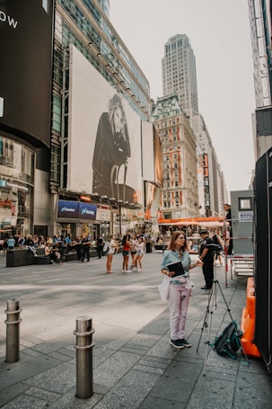 A bustling city street with people walking and gathering. Large billboards and tall buildings fill the scene, with one billboard featuring a monochrome image of a person sitting. A woman stands near a tripod with a bag and a notebook. The street scene is busy with various groups of people interacting and some individuals standing alone.