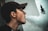 A person wearing a cap is exhaling a large cloud of vapor against a light-colored wall, with part of a distant building visible and slightly out of focus in the background.