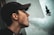 A person wearing a cap is exhaling a large cloud of vapor against a light-colored wall, with part of a distant building visible and slightly out of focus in the background.