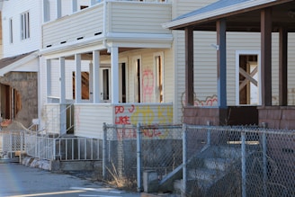 A row of distressed houses with visible signs of wear and graffiti. The windows are either missing or boarded up, and the structures appear to be abandoned. A chain-link fence is in the foreground with various graffiti markings on the siding of the house.