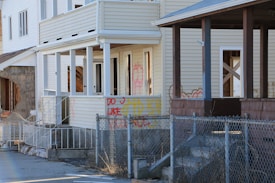 A row of distressed houses with visible signs of wear and graffiti. The windows are either missing or boarded up, and the structures appear to be abandoned. A chain-link fence is in the foreground with various graffiti markings on the siding of the house.