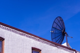 A large satellite dish mounted on a rooftop against a backdrop of clear blue sky. The building features a section of white wall with decorative details near the roofline. The satellite dish is prominent and contrasts with the vivid sky.
