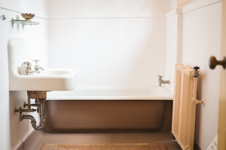 A vintage-style bathroom features an old-fashioned sink with exposed plumbing and cross-handle faucets. A clawfoot bathtub is positioned in the corner, surrounded by plain white walls. A small radiator is affixed to the wall near the bathtub. The floor is covered with a woven rug, adding a touch of warmth to the otherwise stark and minimalistic space.