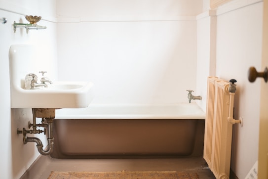 A vintage-style bathroom features an old-fashioned sink with exposed plumbing and cross-handle faucets. A clawfoot bathtub is positioned in the corner, surrounded by plain white walls. A small radiator is affixed to the wall near the bathtub. The floor is covered with a woven rug, adding a touch of warmth to the otherwise stark and minimalistic space.