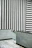 An interior space with minimalist design elements featuring vertical black and white stripes on one wall and horizontal stripes on another. The lower part of the room is lined with light blue and gray tiles, including a small staircase with a metal handrail.