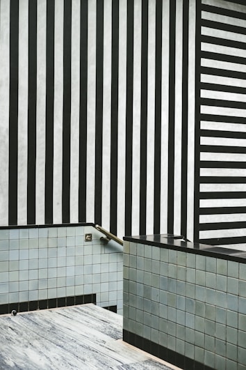 An interior space with minimalist design elements featuring vertical black and white stripes on one wall and horizontal stripes on another. The lower part of the room is lined with light blue and gray tiles, including a small staircase with a metal handrail.