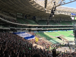 A large stadium with green seating is filled with a crowd of people, many standing and holding a large banner. The roof structure is visible, along with hanging lighting equipment.