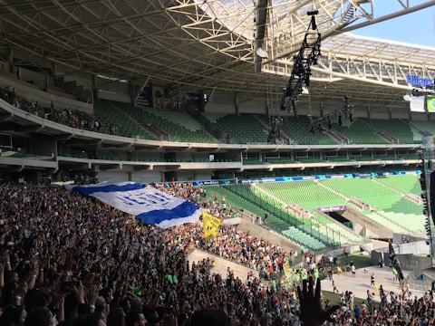 A large stadium with green seating is filled with a crowd of people, many standing and holding a large banner. The roof structure is visible, along with hanging lighting equipment.