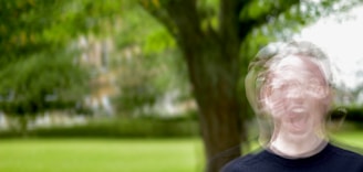 In a blurred outdoor setting, a person with a distorted face appears to be yelling or expressing a strong emotion. The background is dominated by greenery, including a large tree and a grass lawn.