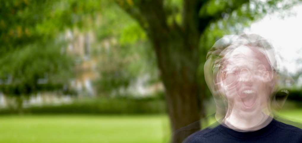 In a blurred outdoor setting, a person with a distorted face appears to be yelling or expressing a strong emotion. The background is dominated by greenery, including a large tree and a grass lawn.