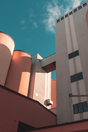 Industrial buildings with large cylindrical silos and rectangular structures connected by an overhead passage, set against a bright sky with a few clouds.