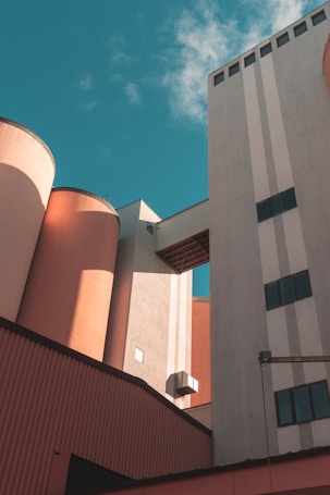 Industrial buildings with large cylindrical silos and rectangular structures connected by an overhead passage, set against a bright sky with a few clouds.