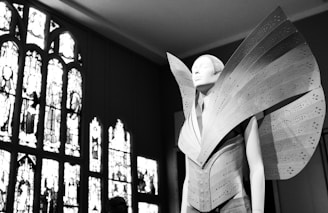 A mannequin adorned with a futuristic and avant-garde garment featuring large, structured, and perforated panels that extend dramatically outward. The setting includes ornate, stained glass windows that provide a contrasting backdrop with intricate designs.