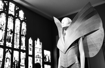 A mannequin adorned with a futuristic and avant-garde garment featuring large, structured, and perforated panels that extend dramatically outward. The setting includes ornate, stained glass windows that provide a contrasting backdrop with intricate designs.