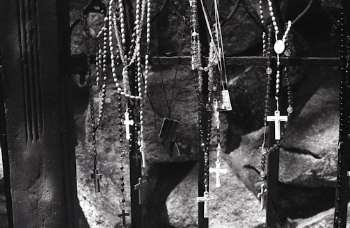 A collection of rosaries with crosses hangs from a dark metal gate. The background consists of rough stone surfaces, suggesting a somber or sacred setting. The image is in black and white, which adds to its solemn atmosphere.