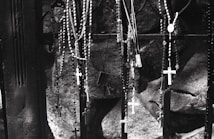 A collection of rosaries with crosses hangs from a dark metal gate. The background consists of rough stone surfaces, suggesting a somber or sacred setting. The image is in black and white, which adds to its solemn atmosphere.