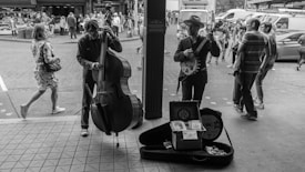 Two street musicians play their instruments in a bustling market area. One musician is playing a double bass, while the other is playing a banjo. A guitar case is open in front of them with tips, and people pass by on the street, suggesting a lively, social atmosphere.