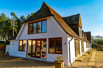 A large, modern house with a white exterior and steep brown roofs. Large windows with wooden frames reflect the surrounding landscape. The building sits on a gravel driveway, bordered by stone pathways and small planters with greenery. In the background, a dense line of tall trees extends under a clear blue sky.