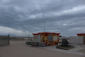 A small, colorful beach hut sits on a sandy beach under a cloudy sky. The hut has vibrant artwork painted on its walls and a sign that reads 'DIREZIO'. Nearby, there are stacks of what appear to be folded beach chairs or loungers, and some netted fences extending across the sand. The overall scene is devoid of people, giving it a serene and slightly desolate atmosphere.
