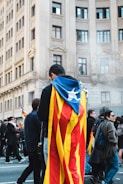 A person wrapped in a flag, likely of Catalonia, stands amidst a group of people on a city street. Tall buildings with classic architecture form the backdrop. The scene appears to be part of a public gathering or demonstration, with several people walking and engaging around.