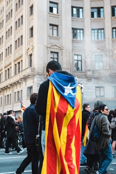 A person wrapped in a flag, likely of Catalonia, stands amidst a group of people on a city street. Tall buildings with classic architecture form the backdrop. The scene appears to be part of a public gathering or demonstration, with several people walking and engaging around.