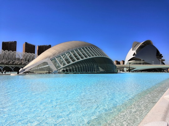Futuristic architectural structures with glass and metal facades sit by a clear blue pool. The buildings feature dome-like curves and sweeping lines, set against a vibrant blue sky. In the background, there are several tall, modern apartments or office buildings.