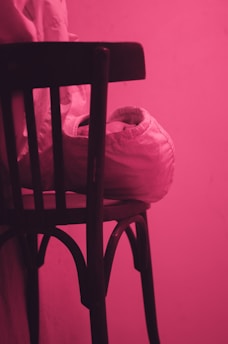 A dark wooden chair with a curved backrest casts soft shadows against a vibrant pink background. Draped over the seat is a partially folded white fabric or jacket, adding contrast to the composition.
