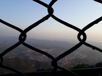 A chain-link fence is in the foreground, with a blurred view of a hilly landscape and a town or city in the background, surrounded by haze under a clear sky.