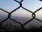 A chain-link fence is in the foreground, with a blurred view of a hilly landscape and a town or city in the background, surrounded by haze under a clear sky.
