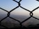 A chain-link fence is in the foreground, with a blurred view of a hilly landscape and a town or city in the background, surrounded by haze under a clear sky.