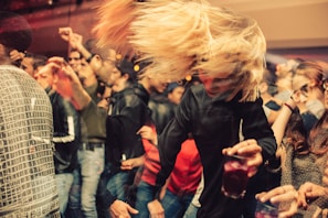 A crowded party scene with people dancing energetically. A person with blonde hair is swinging their head while holding a drink. The room is dimly lit, creating a warm, lively atmosphere.