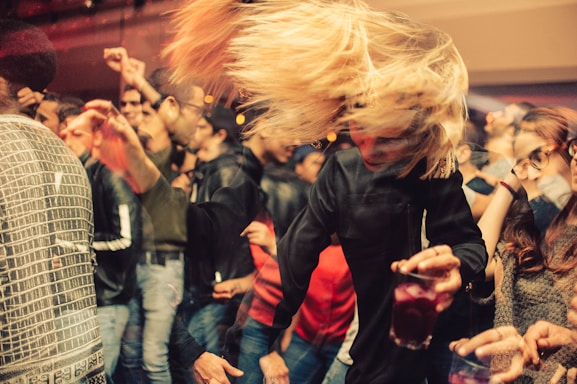 A crowded party scene with people dancing energetically. A person with blonde hair is swinging their head while holding a drink. The room is dimly lit, creating a warm, lively atmosphere.