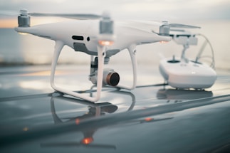 A white quadcopter drone sits on a reflective surface with a remote control device in the background. The background is slightly blurred, suggesting depth of field effects, and the setting appears outdoors during a cloudy day.